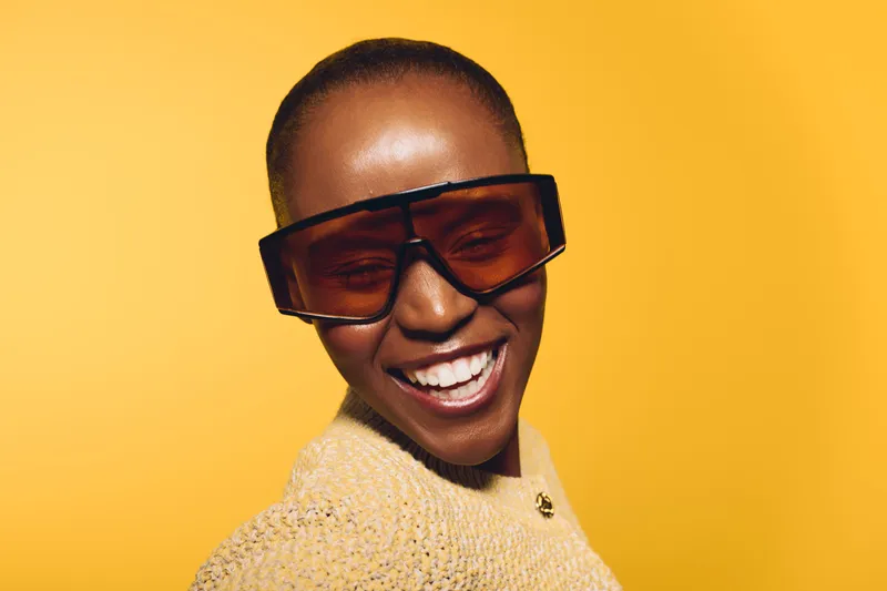 Happy African American woman with dark skin wearing large sunglasses and a yellow sweater smiles against a bright yellow background. Concept of joy, fashion, and positive mood.