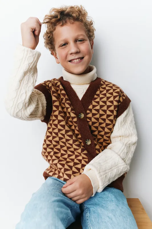 A cheerful young boy sits against a white backdrop, smiling confidently while wearing a geometric patterned knit vest over a cozy cream sweater and light jeans.