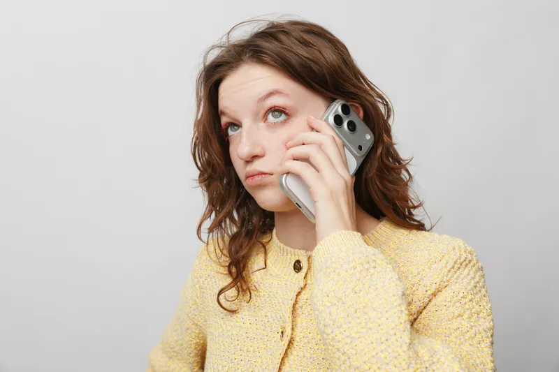 Young woman with curly hair wearing yellow sweater holds smartphone to ear, looking up thoughtfully. Concept of communication, technology, and casual lifestyle in modern portrait.