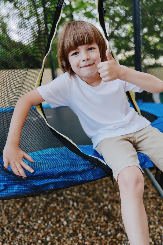 Happy boy giving a thumb up while sitting on a trampoline, wearing a white shirt, enjoying a sunny day outdoors with a playful atmosphere.
