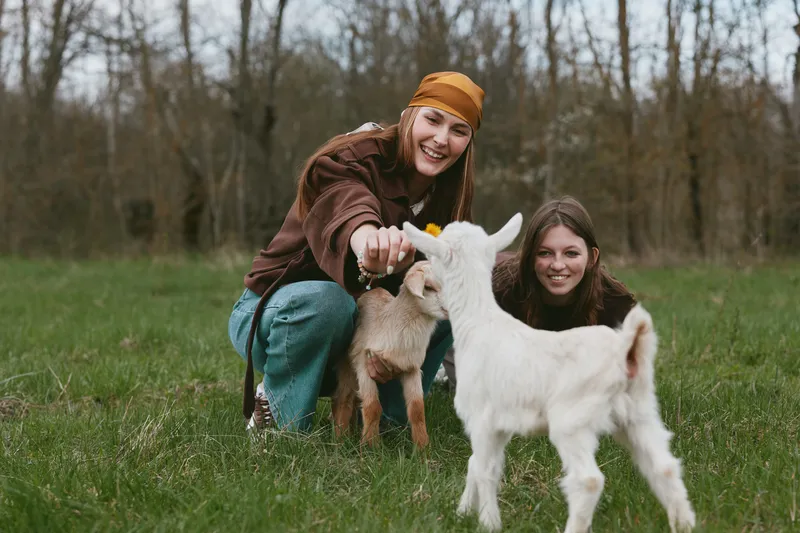 Two smiling women in casual clothing interact with a curious kid goat in a grassy field, capturing a joyful countryside moment and connection between people and animals, shared joy!