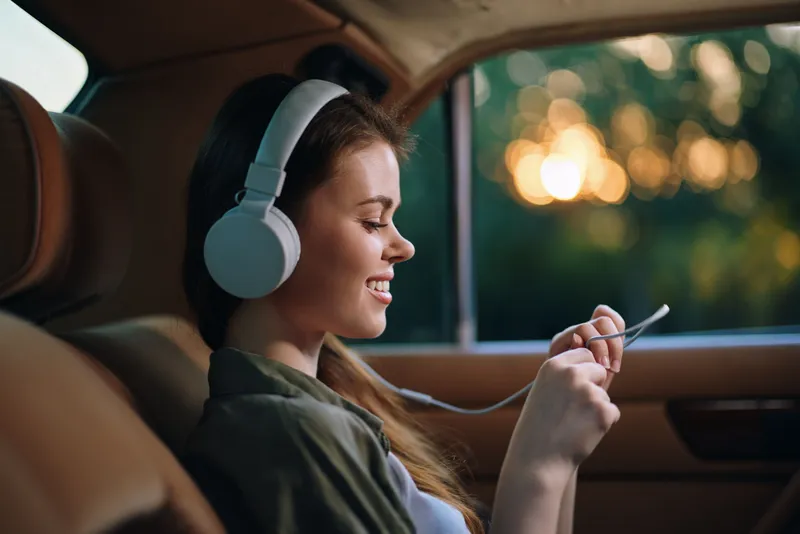 Young woman enjoying music with headphones in a car, sunset background, captures joy and relaxation while using a smartphone.