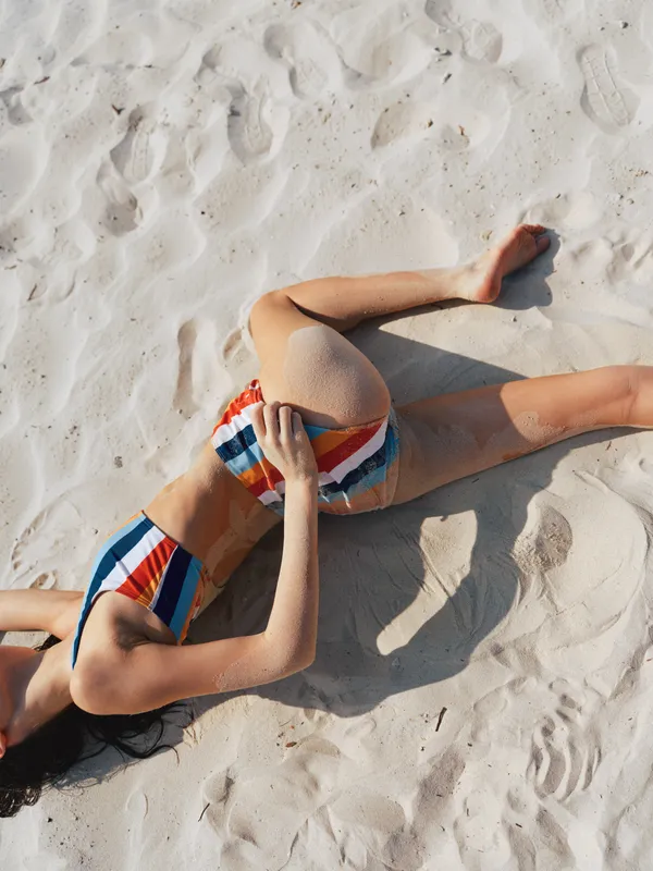 Sunlit beach scene features a person lounging on white sand in a colorful striped bikini, relaxed pose, warm light, soft shadows, and a carefree summer mood.