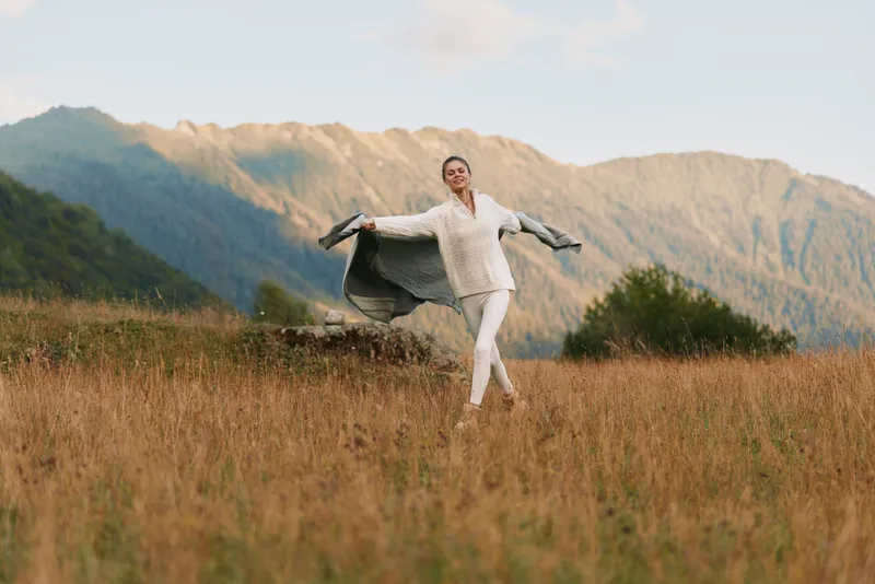 A happy woman in white clothes enjoys freedom and nature while walking through a grassy field with mountains in the background on a clear day.