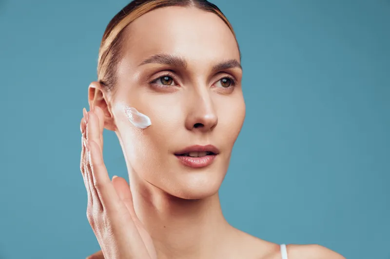 Young woman applying cream on face, skincare routine, clear skin, natural makeup, gentle touch, blue background, beauty concept, closeup portrait, healthy lifestyle