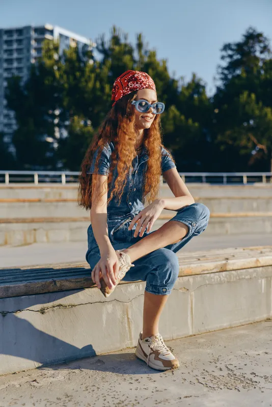 Lifestyle woman in modern boho-western denim street style sitting outdoors, wearing sunglasses and a red bandana with vibrant film color tones on a sunny day.