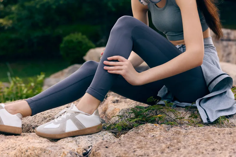 Young woman in sportswear demonstrating healthy lifestyle while sitting outdoors among nature, showcasing athletic wear and relaxed posture during sunny day