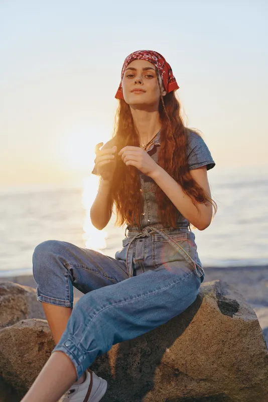 Lifestyle woman in modern boho-western denim street style wearing a red bandana by the sea at sunset with warm film color tones, relaxed and confident atmosphere.