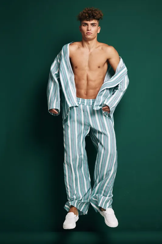 Shirtless young man poses confidently wearing high waist striped loose pants and oversized open matching shirt in bright studio against green background with white sneakers.