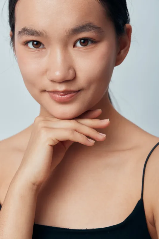 Woman portrait with natural skin and hand under chin wearing black top on light background. Beauty, confidence, youth, and minimal makeup in studio closeup for skincare and wellness.