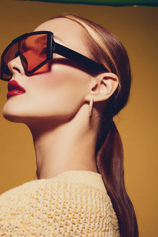Stylish woman with long hair wearing large sunglasses and red lipstick poses against yellow background. Fashion portrait with elegant makeup and modern accessories in warm studio lighting.