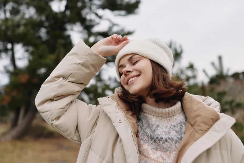 Woman in a warm beige coat and knit sweater, wearing a white beanie, smiling as she enjoys an outdoor winter day with soft natural light and a blurred natural background.