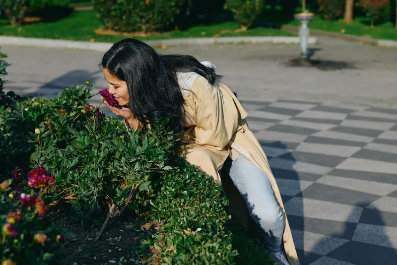 Woman in beige jacket leans toward flowering shrub, smelling blossoms on a garden path, an outdoor moment highlighting nature, casual style and serenity.