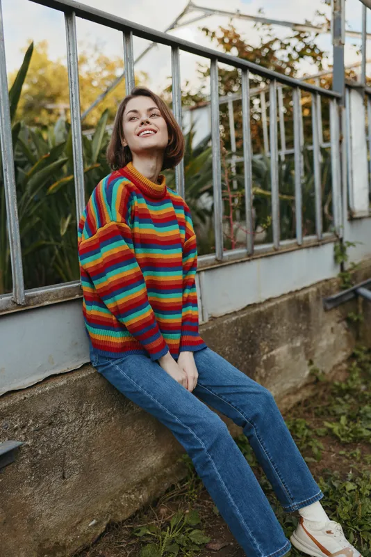 Young woman in a colorful striped sweater smiling while sitting by a glass greenhouse, casual outfit with jeans and white sneakers, looking up relaxed in outdoor garden lifestyle.