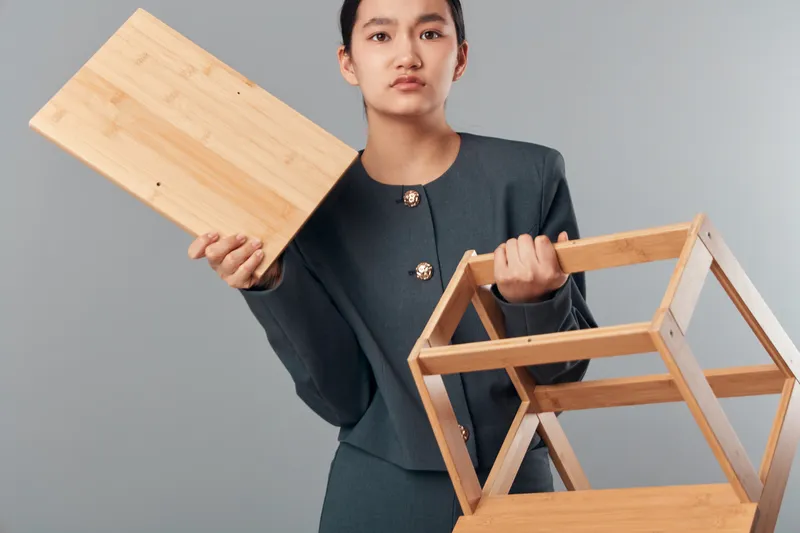 Woman holding wooden shelf and panel in gray outfit standing in studio with neutral background for furniture assembly, interior design, carpentry, and construction tasks in workshop setting.