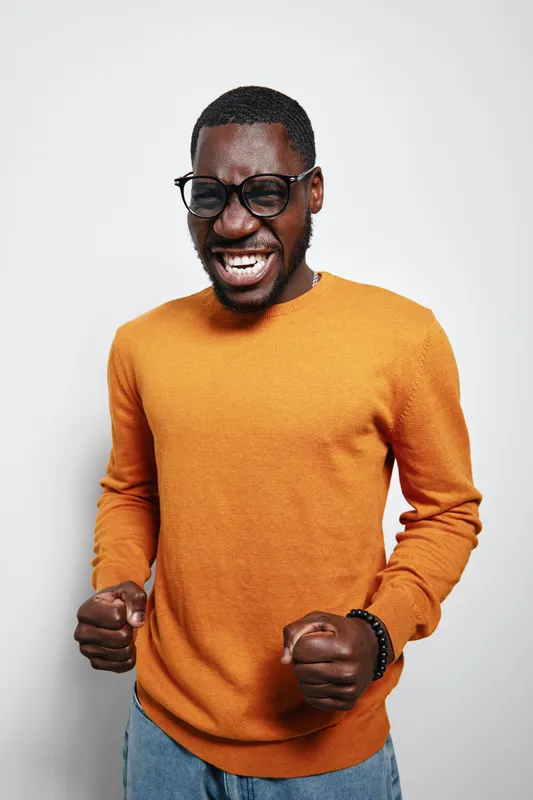 African American man with dark skin wearing glasses and orange sweater expressing excitement with clenched fists and closed eyes on white background. Casual style and joyful emotion.