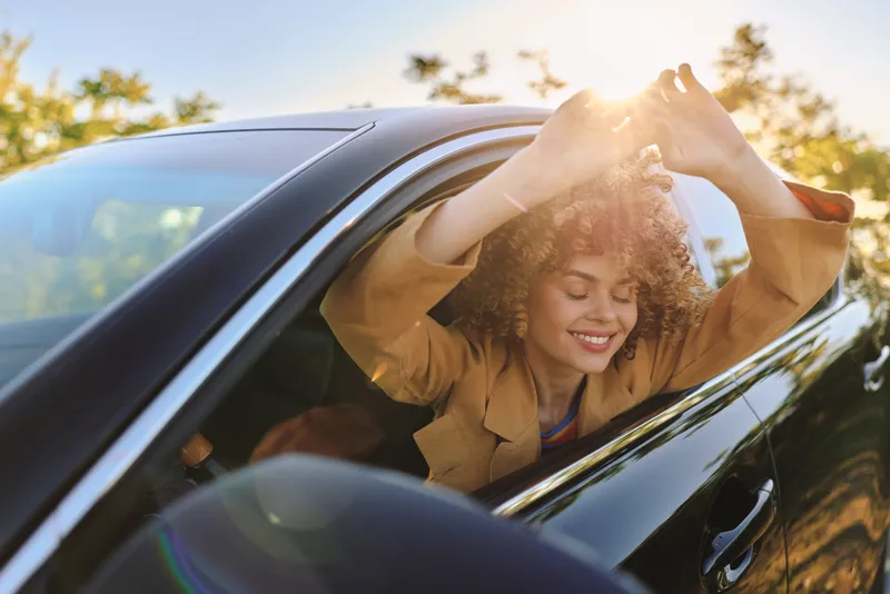 Smiling woman leaning out of a car window in sunlight, a warm outdoors lifestyle moment with travel vibes, happiness and nature foregrounded in a retro color palette.