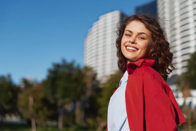 Young woman with curly hair smiles happily while wearing a red jacket outdoors near modern buildings on a sunny day.