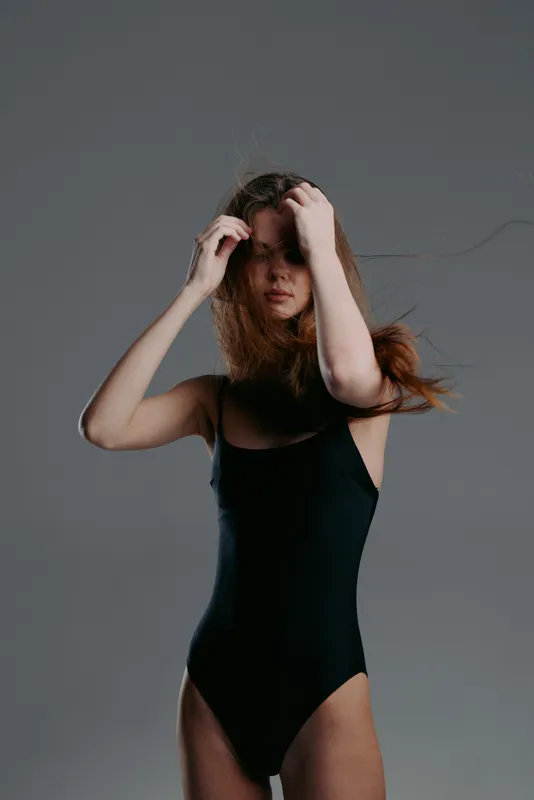 Young woman in black swimsuit posing with loose hair against dark gray background, stylish and moody portrait.