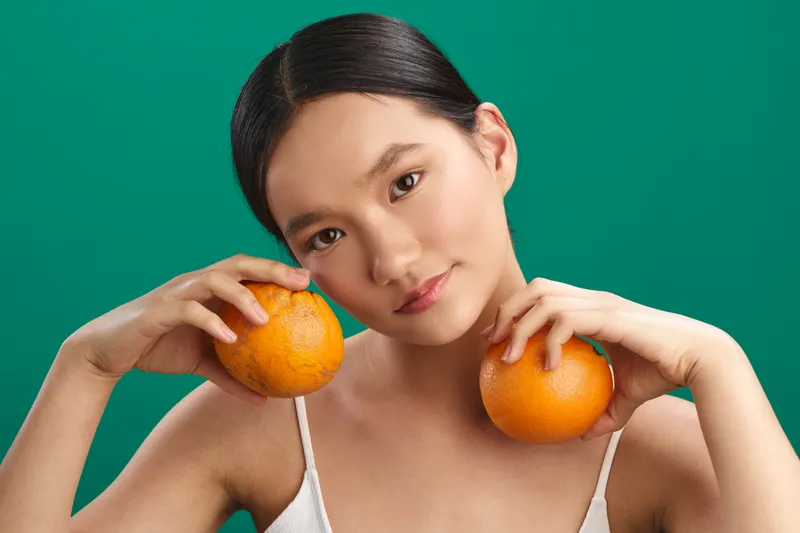 asian woman holds oranges near her cheeks against a bright green background, creating a fresh, playful still life that emphasizes color, health, and a welcoming mood.