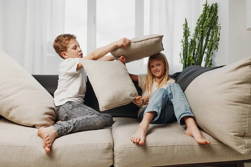 Two playful children having a pillow fight on a beige sofa in a bright living room with casual clothes and natural light.
