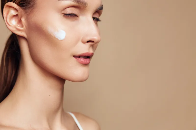 Young woman with fair skin applies white face cream on her cheek while closing her eyes against a beige background, demonstrating a skincare routine and self-care moment.