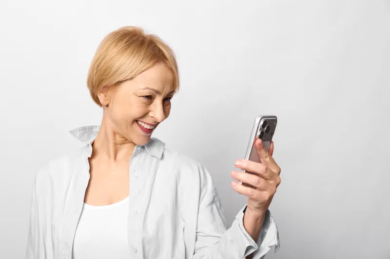 Smiling senior woman in white shirt holds a smartphone, blonde hair, simple bright background, joyful communication with technology, portrait, closeup relaxed expression