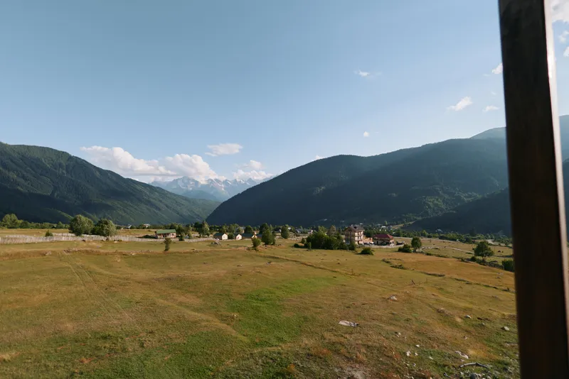A serene valley landscape unfolds with rolling meadow, distant mountains, and scattered houses under a clear blue sky, inviting calm, exploration, and reflection.