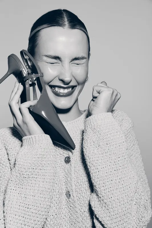 Smiling caucasian woman with fair skin holds red high heel shoe near face, wearing knitted sweater. Concept of joy, fashion and happiness.