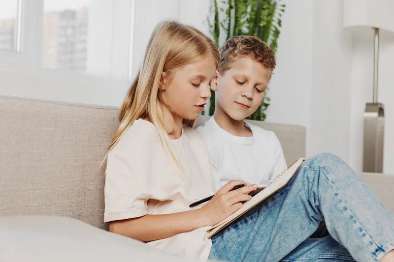 Young siblings sitting on couch drawing together in a bright living room, focused on creativity and family bonding with casual clothes and relaxed atmosphere.