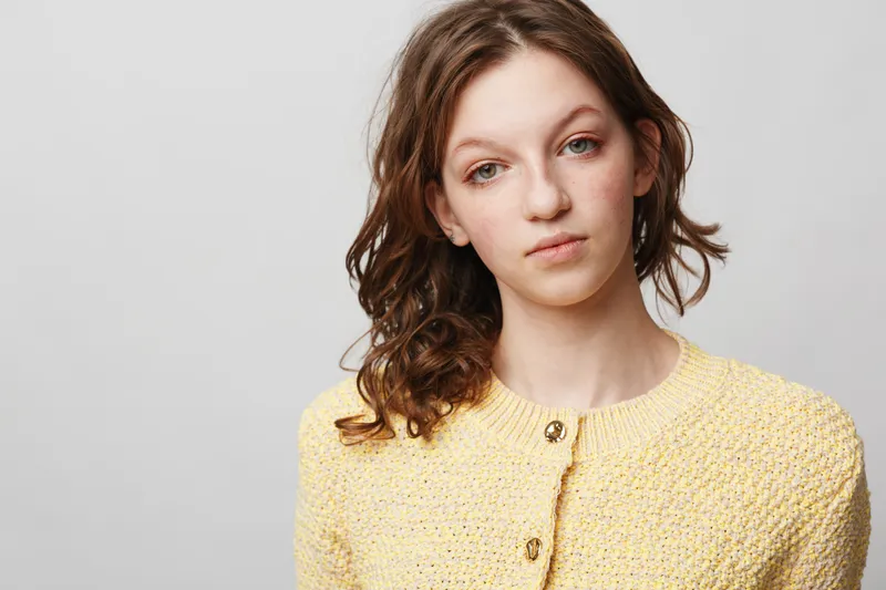 Young woman with curly brown hair wearing yellow knitted sweater looks thoughtfully at camera on plain light background. Concept of contemplation, youth, and natural beauty.