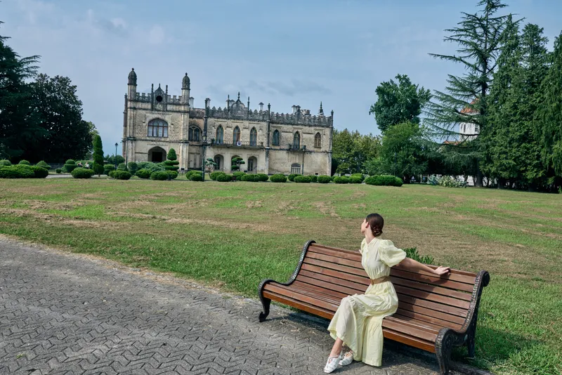 Woman sitting on a bench in a park with a historic castle in the background, wearing a light dress, enjoying a peaceful outdoor moment on a sunny day.
