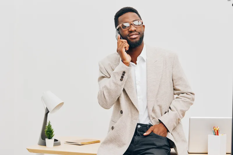 Confident man with glasses in beige blazer and white shirt talking on mobile in modern office setting, looking thoughtful, professional, casual, workspace, business attire, lifestyle concept