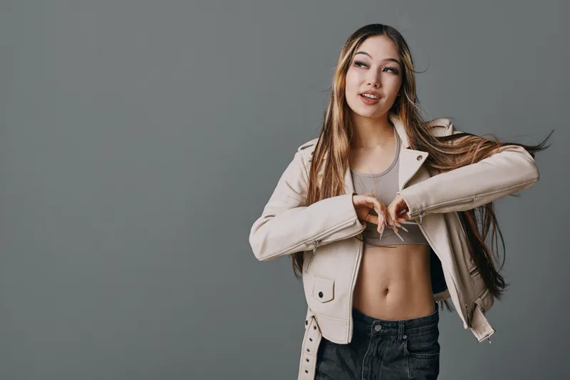 Woman with long hair wearing casual jacket and crop top posing with hands forming heart shape on gray background. Youthful female showing gesture and smiling gently indoors.