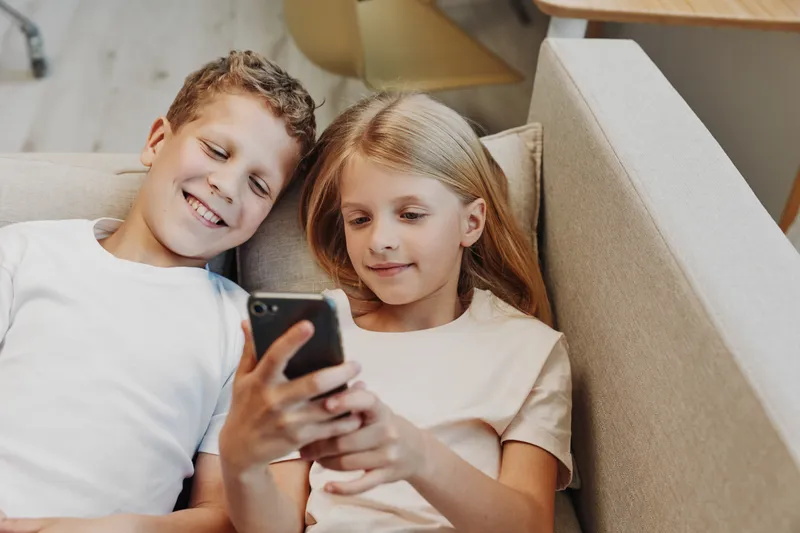 Two children lying on a couch looking at a smartphone screen together. The boy and girl share a relaxed moment in a bright living room. Concept of childhood, technology, and friendship.