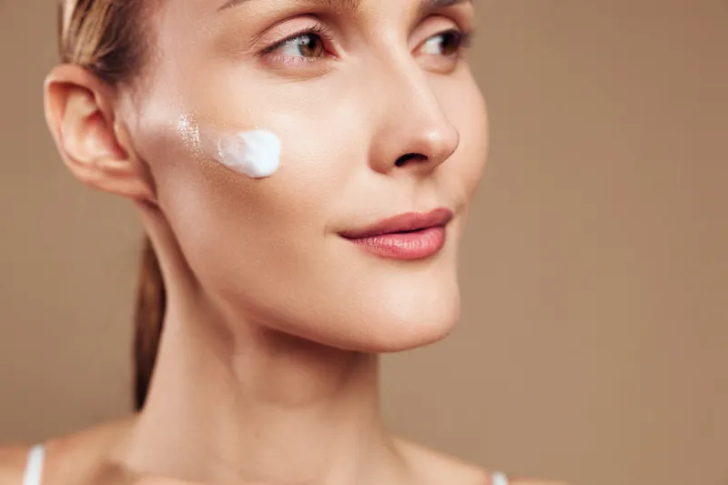 Young woman with light makeup applies cream to her cheek in a close up shot, conveying skincare routine, self care and a calm, focused mood.