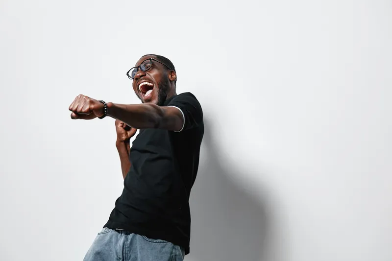 African American man with dark skin wearing glasses and black t shirt posing with energetic punch gesture and joyful expression on white background in studio.