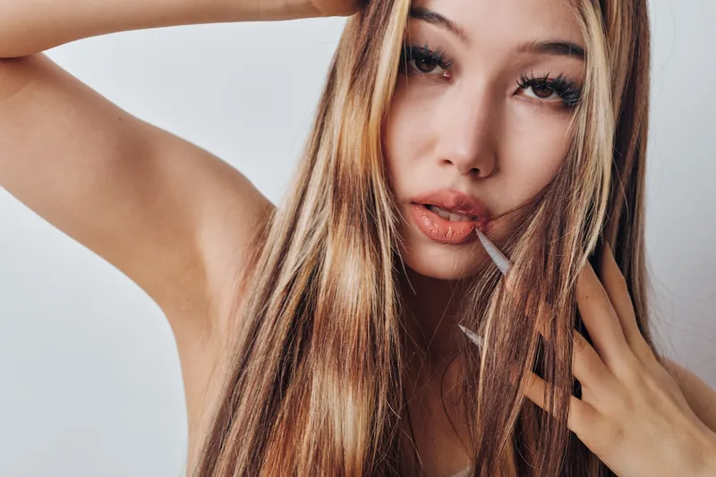 Woman with long blonde hair posing with hand near face and arm raised against white background, makeup and natural expression for beauty and fashion portrait.