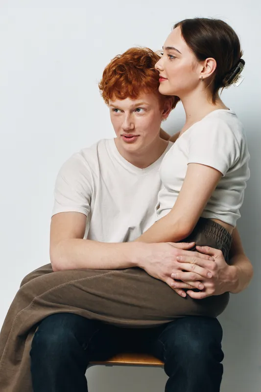 Young couple sitting with woman on man’s lap, casual clothing, intimate moment, love and affection, studio portrait with light background and relaxed expressions.