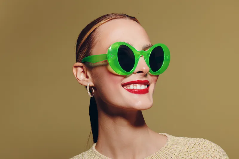 Smiling woman with bright red lipstick wears large green sunglasses and beige sweater. Studio portrait on neutral background. Concept of fashion, style, and confident female model.