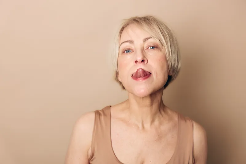 Blonde middle aged woman with short hair sticks out tongue against beige background. Portrait of playful adult female showing expression with casual outfit and natural lighting.
