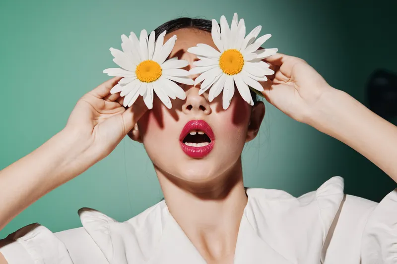 Creative fashion portrait of a young woman holding two large daisies over eyes. Lipstick shines brightly with fresh makeup on turquoise background. Beauty and floral concept.