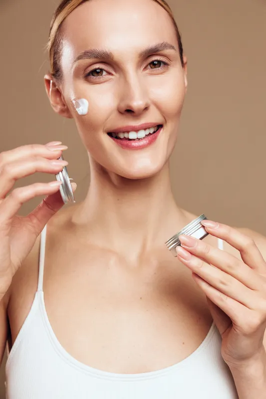 Happy woman applies skincare cream from a jar, smiling, close up portrait, routine beauty moment, clear skin, isolated on soft beige background, hands applying cream, gentle glow, healthy complexion