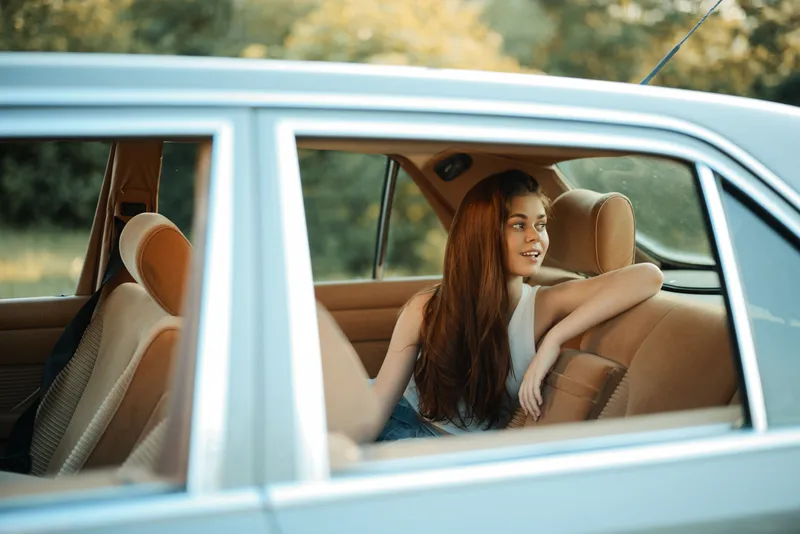 Young woman with long hair sitting in a vintage car, enjoying a joyful moment in a natural setting, wearing a casual outfit, conveying relaxation and happiness.