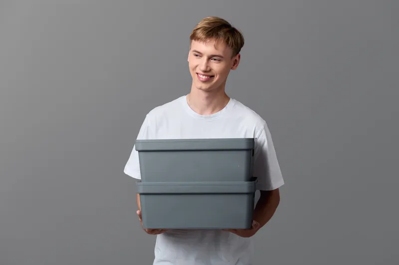 Casual man holding two grey storage boxes in a plain studio backdrop, showcasing organized packaging and home storage ideas with a clean, minimalist vibe.