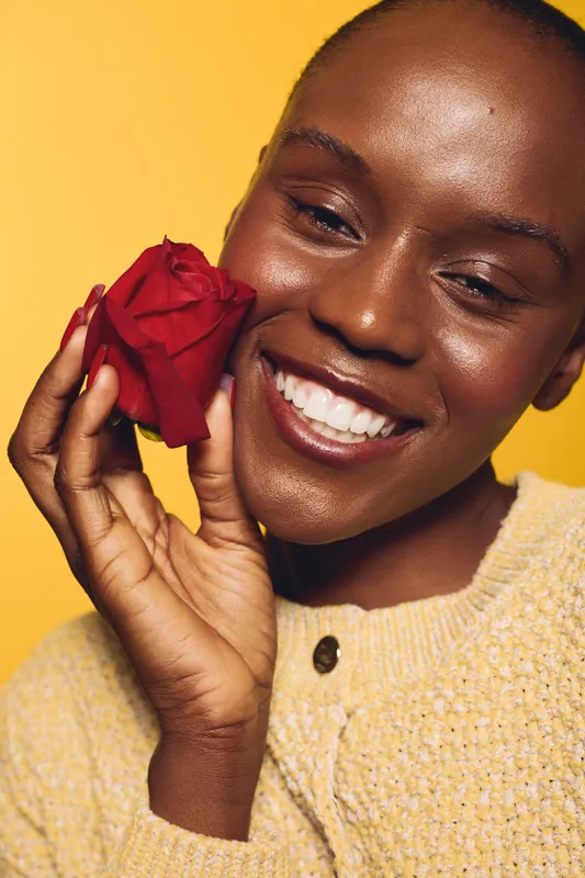 Smiling African woman with dark skin holds red rose near face, wearing beige sweater. Bright yellow background creates warm mood. Concept of happiness, beauty, floral, and natural skin care.