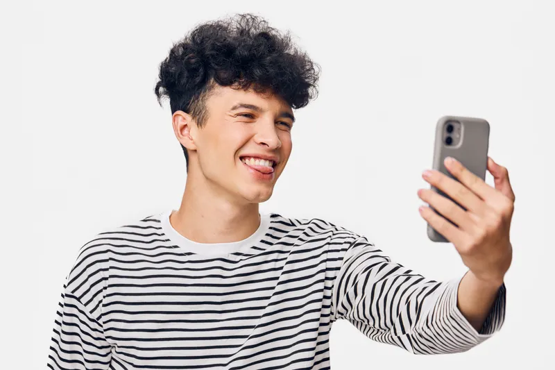 Young man with curly hair taking a selfie with a smartphone, wearing a casual striped shirt, smiling playfully against a plain white background.