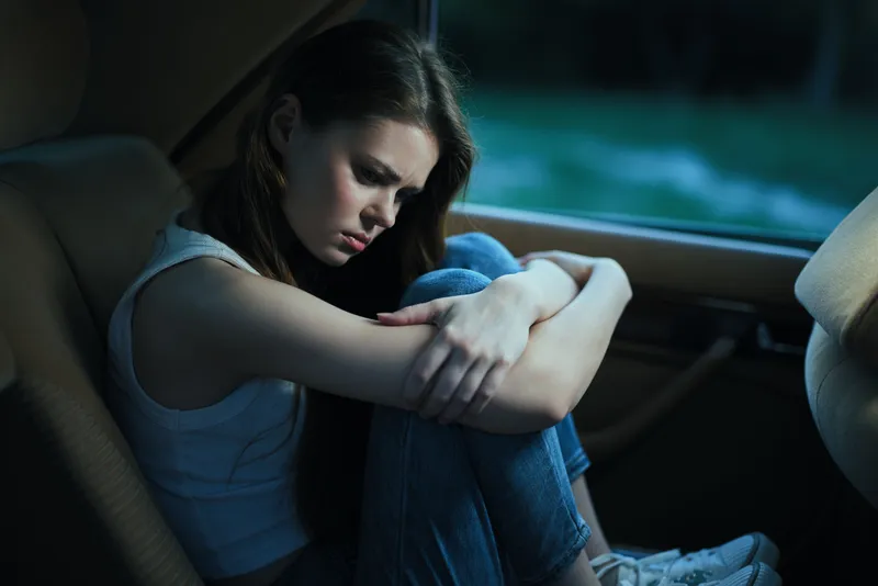 A young woman of Caucasian descent sits in a car, showing emotions of sadness and reflection while embracing her knees in a dimly lit environment.