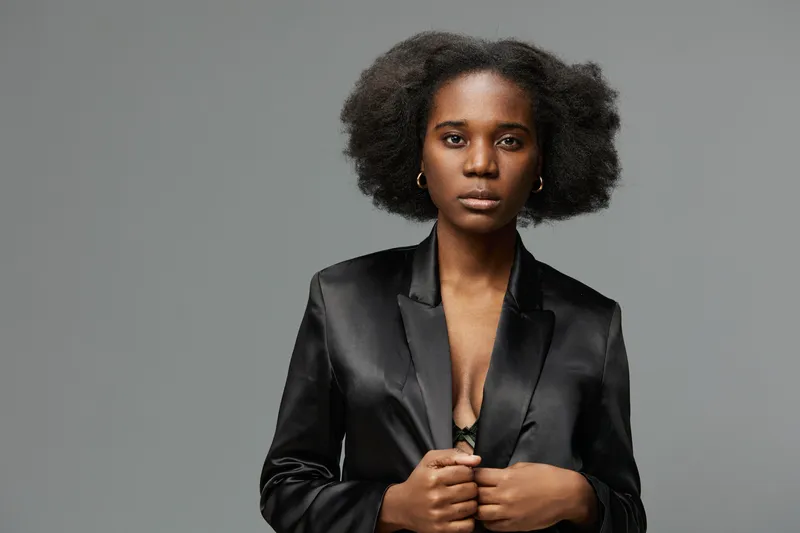 Confident Black woman in a stylish black blazer, showcasing natural hair, set against a minimalist gray background, exuding strength and sophistication