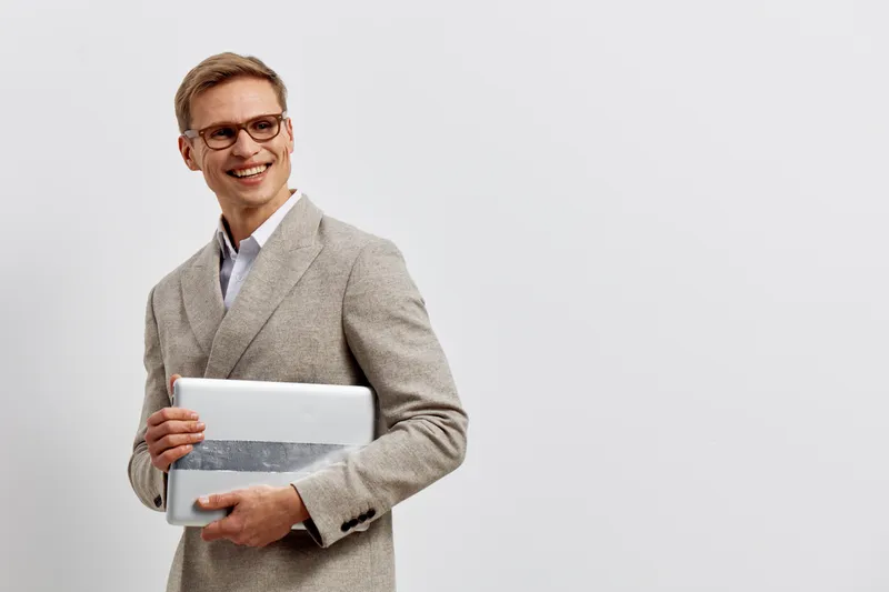 Smiling professional man in beige blazer holding a silver laptop in hand on plain white background, studio portrait. Business casual, confident, cheerful, mature, confident businessman concept.